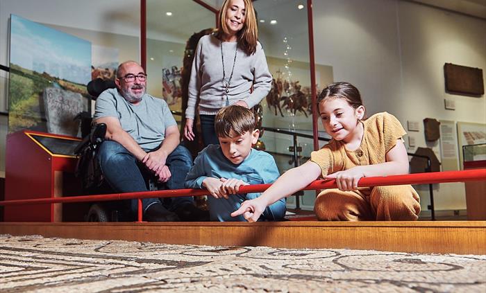 a young girl and boy, with mum and dad are in the museum gallery. the children are crouched down looking at a large Roman mosaic on display at floor l